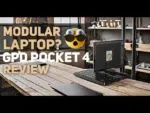 Slika s modularnim prijenosnim računalom, GPD Pocket 4, na modernom drvenom stolu u radnom prostoru u industrijskom stilu. Na preklapanju teksta piše 'Modularno prijenosno računalo? GPD Pocket 4 recenzija' s izražajnim emotikonom koji prikazuje iznenađenje, ističući jedinstveni modularni dizajn uređaja.