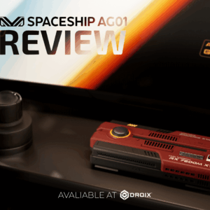 A product review image features a dark red rectangular device, possibly a graphics card or mini PC, with "RX 7600M XT" and "AYANEO GRAPHICS STARS" visible on it. In the background, a screen displays "SPACESHIP AG01 REVIEW" with a logo, and below the device, text reads "AVALIABLE AT DROIX." A small, dark, cylindrical object is to the left of the device.