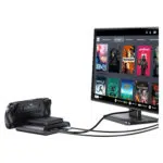 Črna ročna konzola GPD WIN 5 je prikazana priklopljena in s kabli povezana z monitorjem. Na monitorju je prikazan vmesnik v slogu sistema Steam OS z mrežo igralnih naslovov, kot so Dead Cells, Deathloop, Portal 2 in Stardew Valley, kar ponazarja zmožnost naprave, da poganja namizno igralno izkušnjo. 